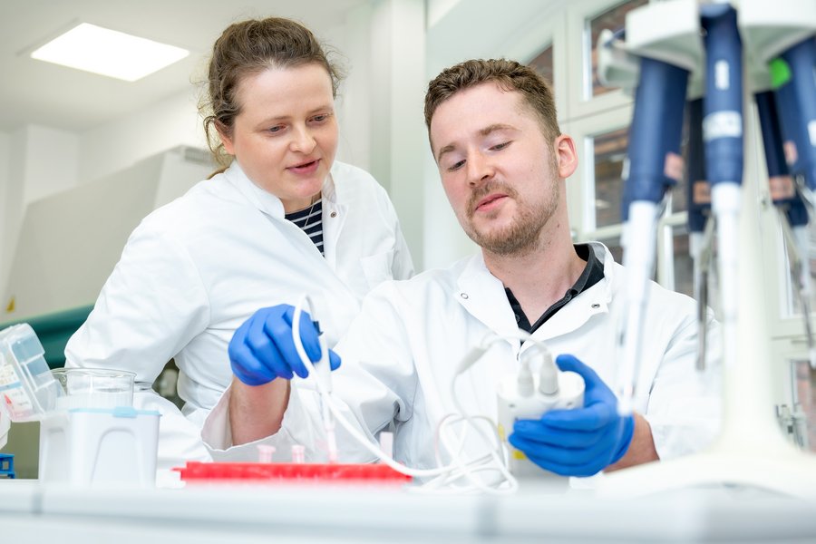 Dr. Stefanie Ruhs und Philipp Terpe arbeiten mit einem technischen Gerät im Labor. Im Vordergrund ist ein Ständer mit mehreren Pipetten unscharf zu erkennen.  Dr. Stefanie Ruhs und Philipp Terpe arbeiten mit einem technischen Gerät im Labor. Im Vordergrund ist ein Ständer mit mehreren Pipetten unscharf zu erkennen.