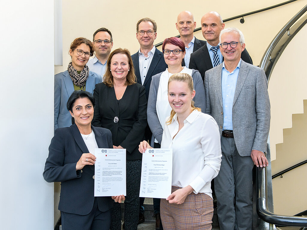 Zehn Personen stehen gestaffelt auf einer geschwungenen Treppe und lächeln in die Kamera. Zwei Frauen in der ersten Reihe halten Urkunden.