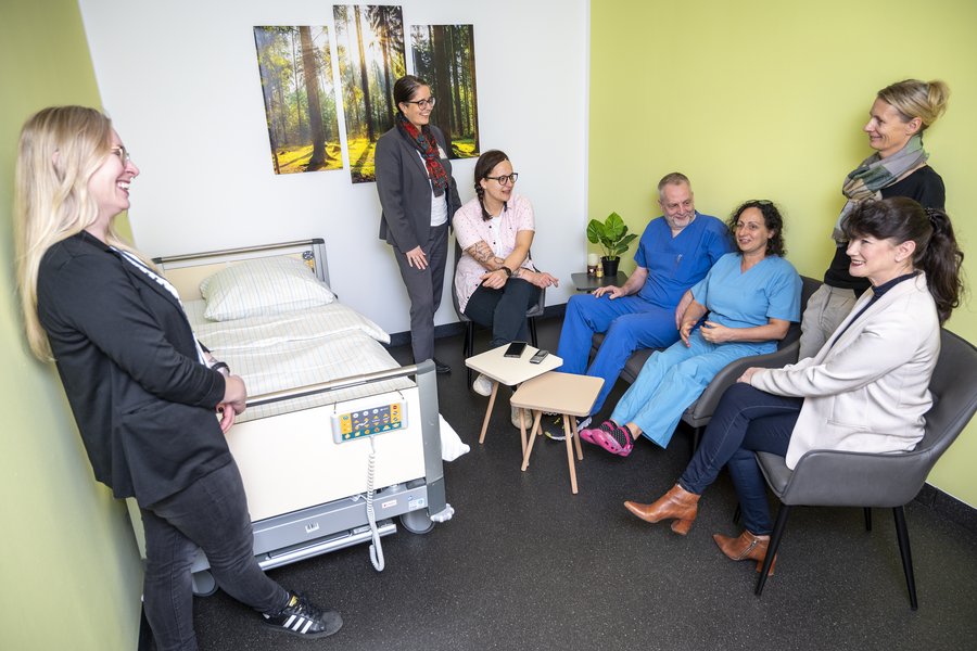 Eine Gruppe von Mitarbeitenden befindet sich im neuen Hybriden Abschiedsraum des Universitätsklinikums Halle (Saale). Einige Personen stehen, einige sitzen. Links an der Wand steht ein Krankenhausbett, rechts ein Sofa und Sessel. An der Wand hängt ein Bild mit Waldmotiv.  Eine Gruppe von Mitarbeitenden befindet sich im neuen Hybriden Abschiedsraum des Universitätsklinikums Halle (Saale). Einige Personen stehen, einige sitzen. Links an der Wand steht ein Krankenhausbett, rechts ein Sofa und Sessel. An der Wand hängt ein Bild mit Waldmotiv.