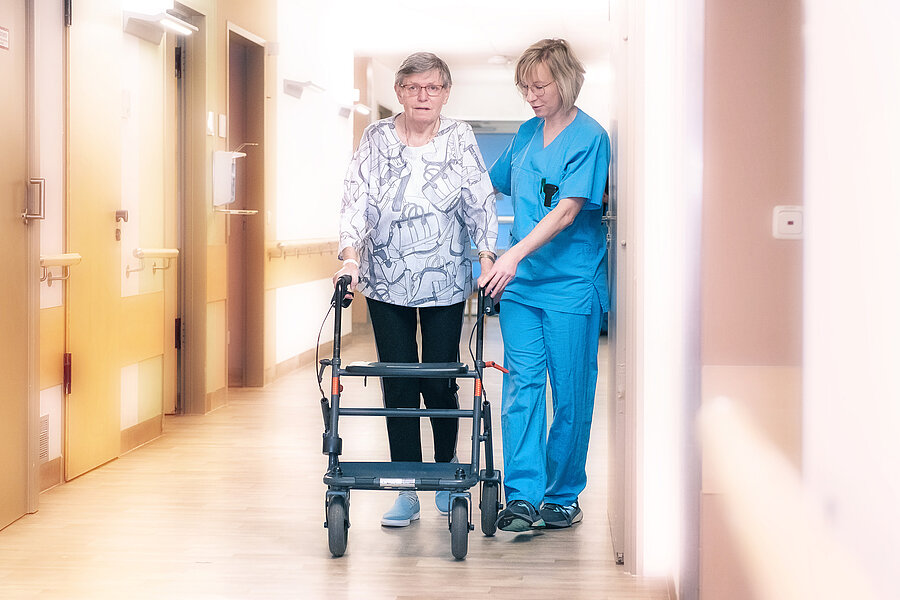 Eine ältere Dame und eine Person in medizinischer Arbeitskleidung gehen einen Krankenhausflur entlang. Die ältere Dame nutzt einen Rollator, die andere Person unterstützt sie.  Eine ältere Dame und eine Person in medizinischer Arbeitskleidung gehen einen Krankenhausflur entlang. Die ältere Dame nutzt einen Rollator, die andere Person unterstützt sie.