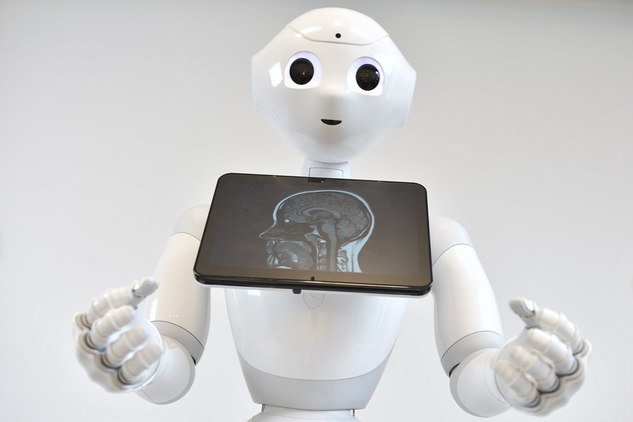 Weißer Roboter in der Größe eines Kindes zeigt auf einem Monitor die Abbildung einer MRT-Aufnahme des Kopfes.  Weißer Roboter in der Größe eines Kindes zeigt auf einem Monitor die Abbildung einer MRT-Aufnahme des Kopfes.