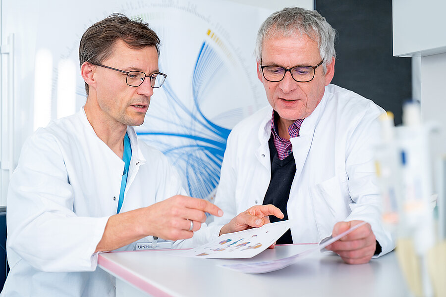 Two men in white lab coats are sitting at a table looking at paperwork. In the background, there is a circular graphic with many lines, and in the foreground, there are blurred test tubes. Two men in white lab coats are sitting at a table looking at paperwork. In the background, there is a circular graphic with many lines, and in the foreground, there are blurred test tubes.