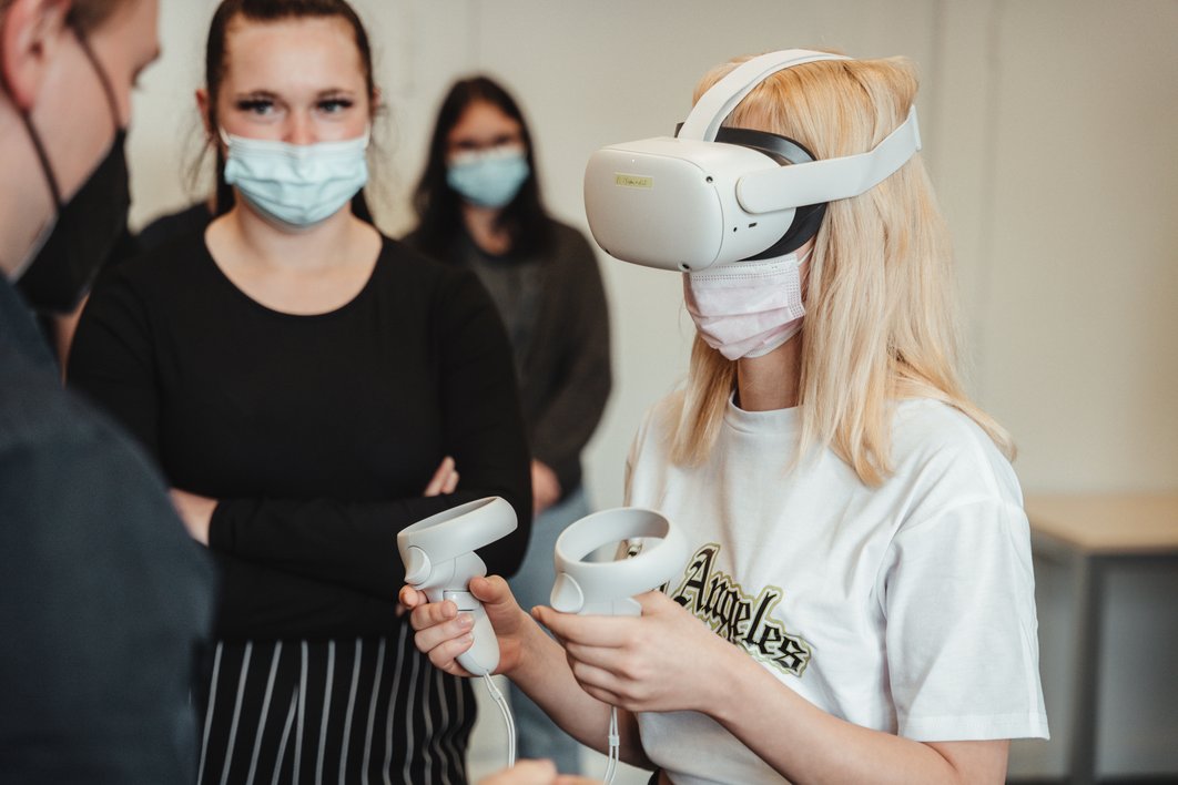 Frau mit VR-Brille und Joysticks in der Hand, drei weitere im Raum