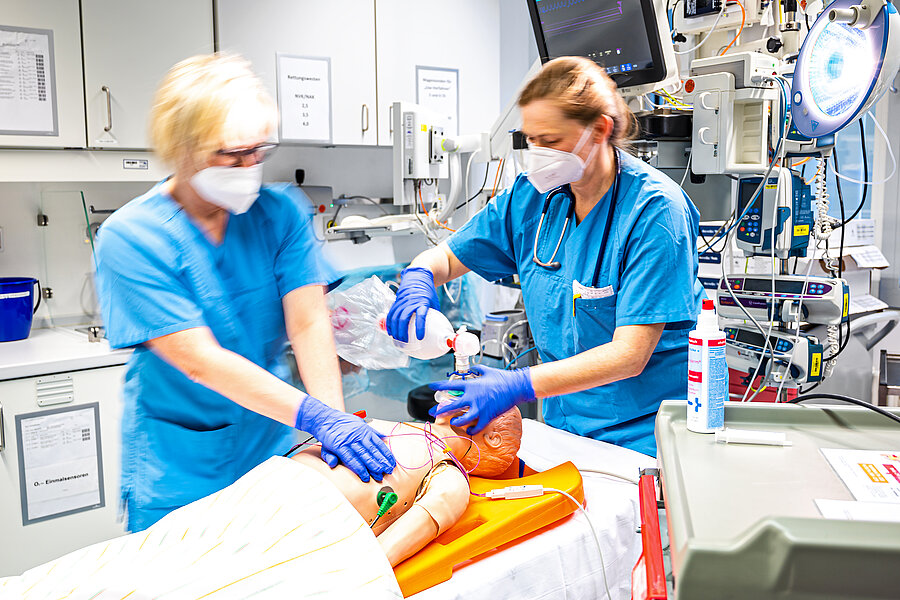 Zwei Personen in medizinischer Arbeitskleidung stehen in einem Behandlungsraum eines Krankenhauses. Beide tragen Masken und Handschuhe und trainieren eine Notfallsituation anhand einer Simulationspuppe. Im Hintergrund befinden sich Schränke und medizinische Geräte.  Zwei Personen in medizinischer Arbeitskleidung stehen in einem Behandlungsraum eines Krankenhauses. Beide tragen Masken und Handschuhe und trainieren eine Notfallsituation anhand einer Simulationspuppe. Im Hintergrund befinden sich Schränke und medizinische Geräte.