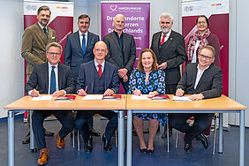 Gruppenfoto einer Vertragsunterzeichnung: Vier Personen sitzen an zwei Tischen und unterzeichnen Dokumente, hinter ihnen stehen weitere Beteiligte. Im Hintergrund sind Roll-ups des Universitätsklinikums Halle (UKH) und des Harzklinikums zu sehen