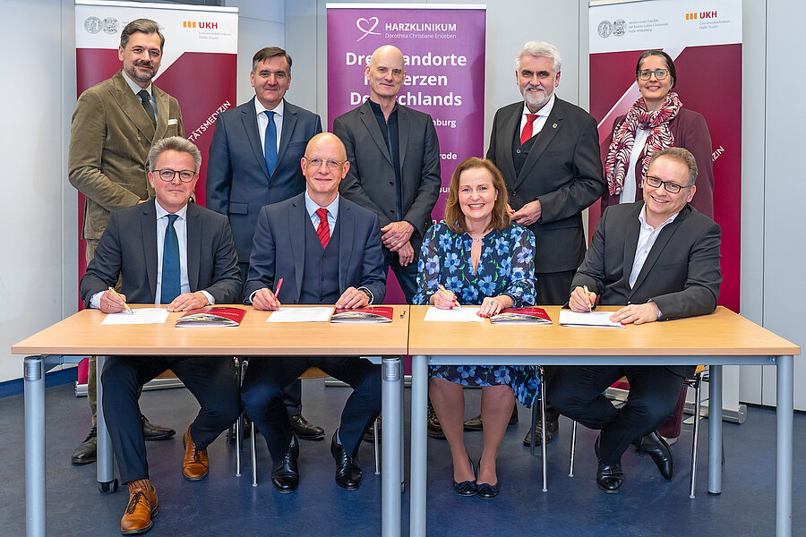 Gruppenfoto einer Vertragsunterzeichnung: Vier Personen sitzen an zwei Tischen und unterzeichnen Dokumente, hinter ihnen stehen weitere Beteiligte. Im Hintergrund sind Roll-ups des Universitätsklinikums Halle (UKH) und des Harzklinikums zu sehen  Gruppenfoto einer Vertragsunterzeichnung: Vier Personen sitzen an zwei Tischen und unterzeichnen Dokumente, hinter ihnen stehen weitere Beteiligte. Im Hintergrund sind Roll-ups des Universitätsklinikums Halle (UKH) und des Harzklinikums zu sehen