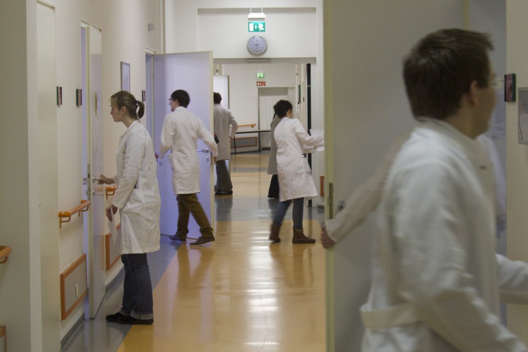 Mehrere Personen in Arztkitteln öffnen in einem Flur Türen oder betreten Zimmer.