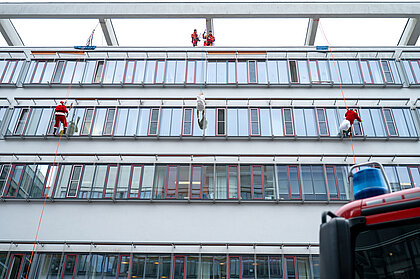 Mehrere verkleidete Höhenretter:innen seilen sich an der Fassade eines Klinikgebäudes ab, während Kolleg:innen vom Dach aus sichern.