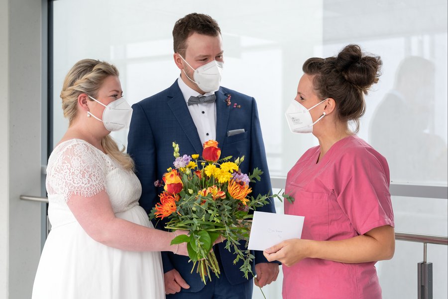 zu sehen ist links eine Braut in Brautkleid, in der Mitte ein Bräutigam im Anzug. Rechts überreicht eine Frau in medizinischer Arbeitskleidung (magenta) einen Blumenstrauß und einen Umschlag.  zu sehen ist links eine Braut in Brautkleid, in der Mitte ein Bräutigam im Anzug. Rechts überreicht eine Frau in medizinischer Arbeitskleidung (magenta) einen Blumenstrauß und einen Umschlag.