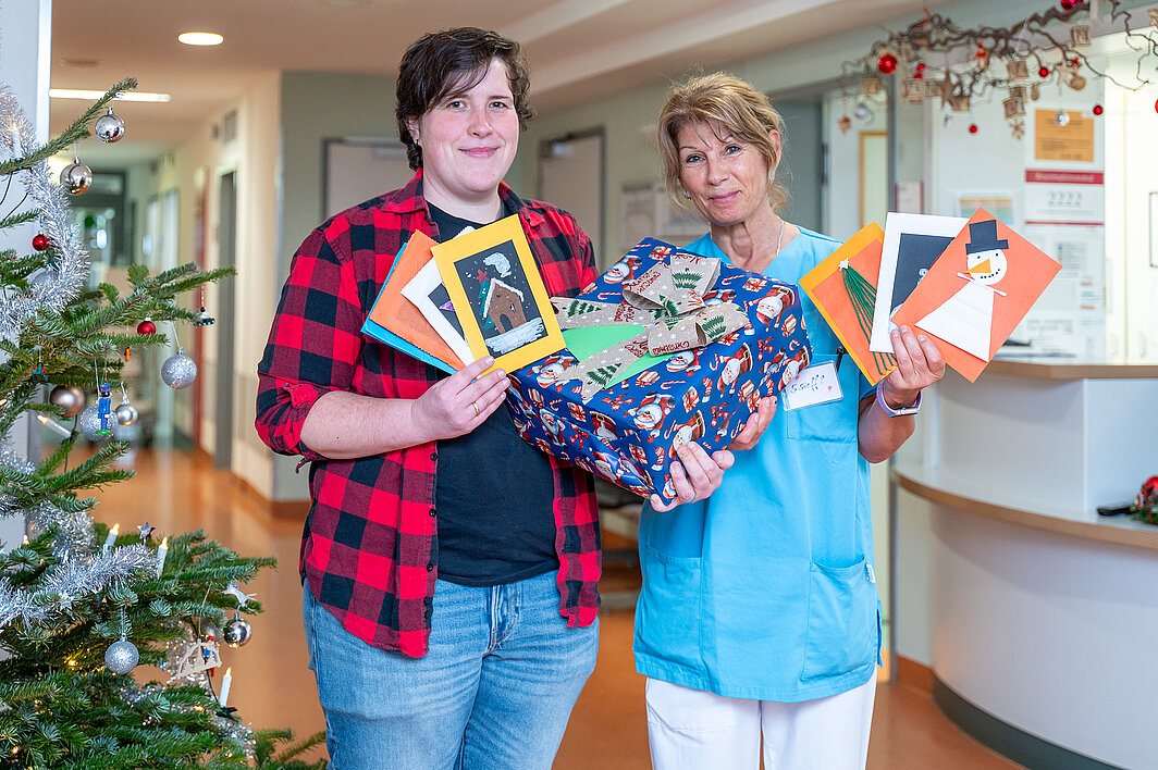 Zwei Personen stehen in einem Krankenhausflur und halten Weihnachtskarten und ein Geschenk in den Händen. Die Person rechts trägt medizinische Arbeitskleidung. Links im Bild steht ein Weihnachtsbaum im Anschnitt.