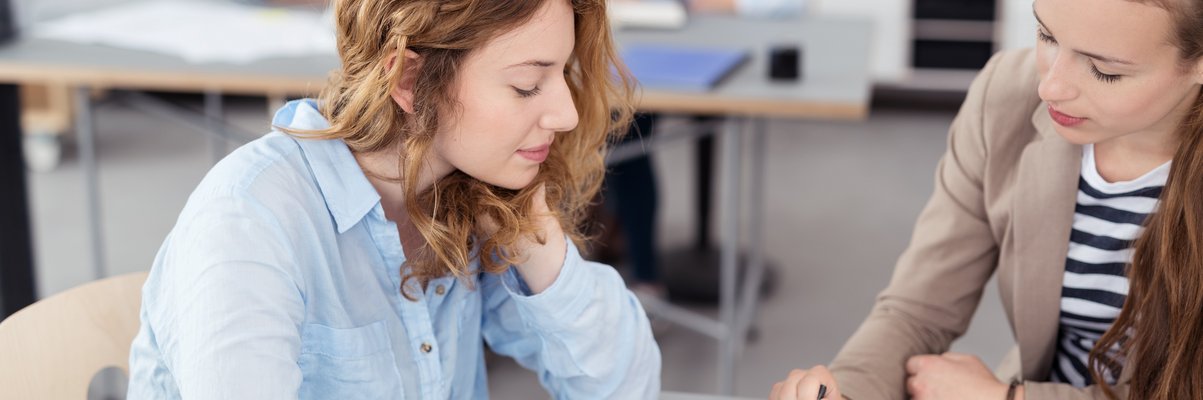 Zwei Frauen im Gespräch an einem Tisch. Auf dem Tisch liegen Blätter. Eine Frau zeigt mit dem Stift auf ein Detail auf dem Blatt. Im Hintergrund sitzen weitere Menschen an einem Tisch