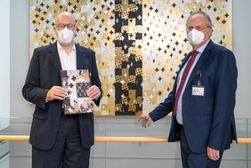 zwei Männer in dunklen Anzügen stehen an einer Brüstung. Im Hintergrund ist eine Wandinstallation in den Farben Schwarz, Weiß und Gold zu sehen. Der Mann links hält ein Buch in den Händen