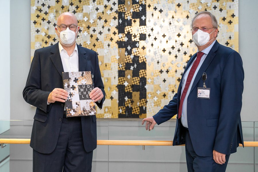 zwei Männer in dunklen Anzügen stehen an einer Brüstung. Im Hintergrund ist eine Wandinstallation in den Farben Schwarz, Weiß und Gold zu sehen. Der Mann links hält ein Buch in den Händen  zwei Männer in dunklen Anzügen stehen an einer Brüstung. Im Hintergrund ist eine Wandinstallation in den Farben Schwarz, Weiß und Gold zu sehen. Der Mann links hält ein Buch in den Händen