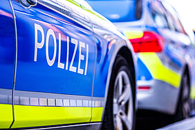 Aus der Froschperspektive sind zwei Polizeiautos aufgenommen. Im Fokus steht die auffällige Folierung des Fahrzeugs im Bildvordergrund in Polizeifarben und der Schriftzug Polizei.