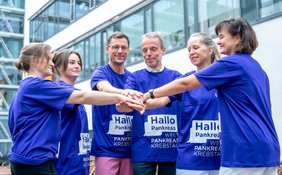 Sechs Personen stehen vor einem Gebäude im Halbkreis und halten jeweils einen Arm in die Mitte, sodass die Hände übereinanderliegen. Alle tragen Motto-T-Shirts zum Weltpankreaskrebstag.