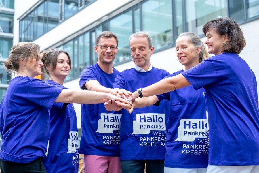 Sechs Personen stehen vor einem Gebäude im Halbkreis und halten jeweils einen Arm in die Mitte, sodass die Hände übereinanderliegen. Alle tragen Motto-T-Shirts zum Weltpankreaskrebstag.  Sechs Personen stehen vor einem Gebäude im Halbkreis und halten jeweils einen Arm in die Mitte, sodass die Hände übereinanderliegen. Alle tragen Motto-T-Shirts zum Weltpankreaskrebstag.