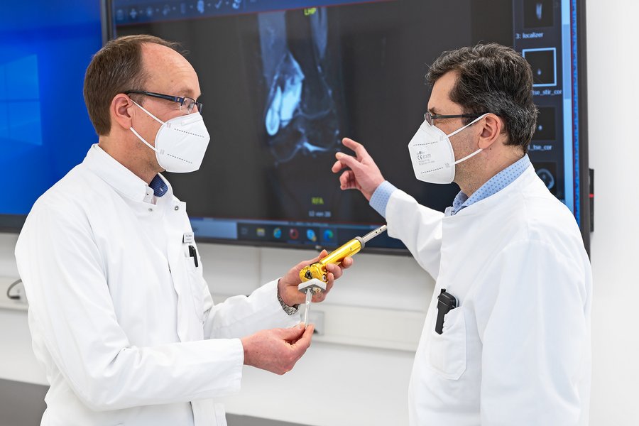 zwei Männer in weißen Arztkittel stehen vor einem Monitor, auf dem ein Röntgenbild zu sehen ist. Der Mann auf der linken Seite hält ein künstliches Kniegelenk in der Hand  zwei Männer in weißen Arztkittel stehen vor einem Monitor, auf dem ein Röntgenbild zu sehen ist. Der Mann auf der linken Seite hält ein künstliches Kniegelenk in der Hand