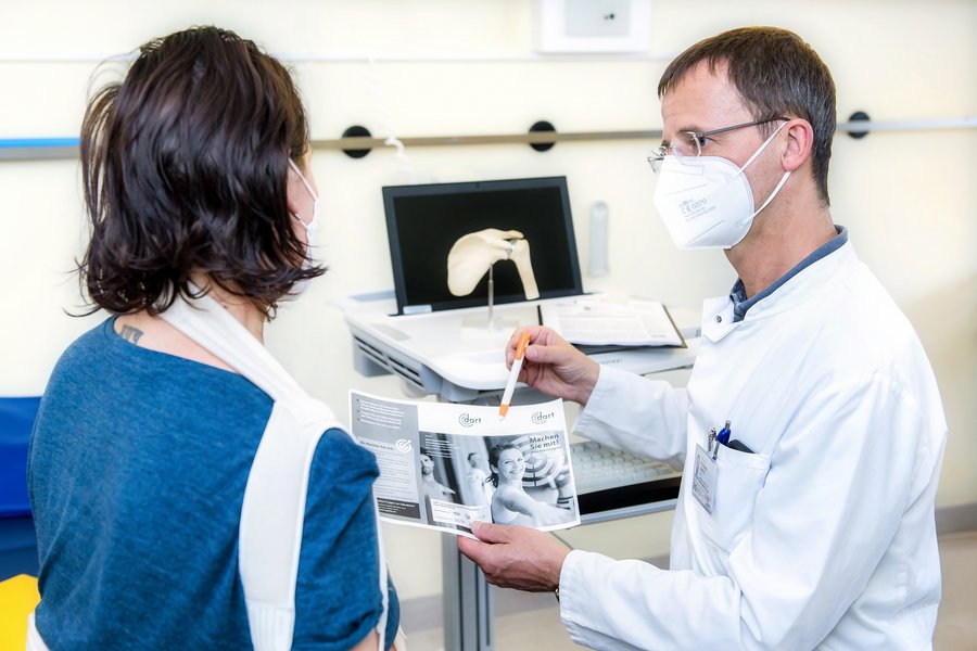 Ein Arzt rechts im Bild unterhält sich mit einer Patientin links von ihm. Ihr rechter Arm ist in einer Schlinge. Der Arzt hat ein Blatt in der Hand auf das er deutet.   Ein Arzt rechts im Bild unterhält sich mit einer Patientin links von ihm. Ihr rechter Arm ist in einer Schlinge. Der Arzt hat ein Blatt in der Hand auf das er deutet. 