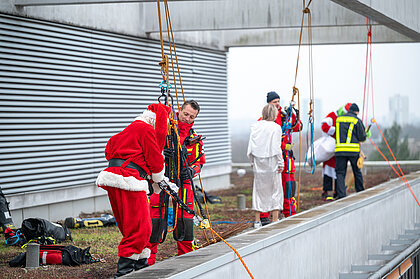 Feuerwehrkräfte sichern auf einem Klinikdach ihre Abseilausrüstung, während drei als Weihnachtsmann, Engel und Grinch verkleidete Höhenretter:innen vorbereitet werden.