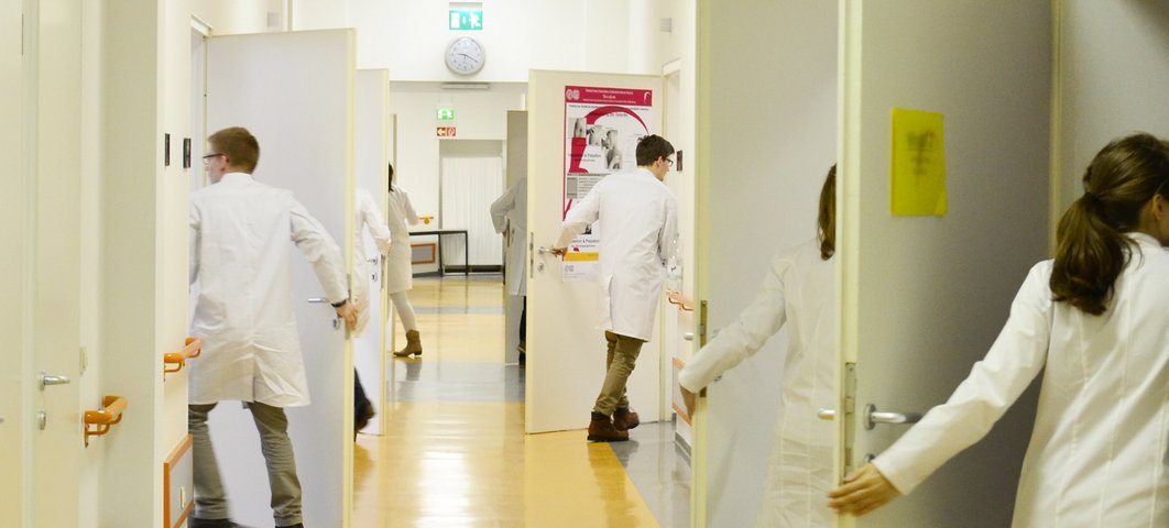 Blick in einen Flur: Mehrere Personen in Arztkitteln betreten zu beiden Seiten des Flures verschiedene Zimmer.
