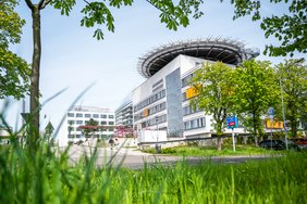 im Hintergrund ist das Hauptgebäude des Universitätsklinikum Halle von schräg vorne zu sehen. Im Vordergrund sieht man grünes Gras und belaubte Bäume rahmen das Beild ein