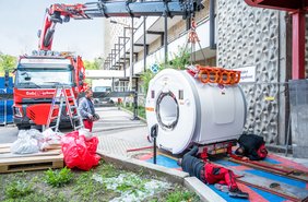 Ein Schwertransporter lädt mit einem Kran eine MRT-Röhre ab. vor der Röhre liegen zwei Arbeiter in roter Arbeitskleidung auf dem Boden und arbeiten. Im Hintergrund ist eine Baustelle zu sehen.