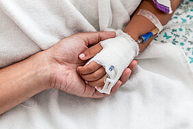 Detailaufnahme: Die Hand eines Kindes liegt in der Hand einer erwachsenen Person. In der Hand des Kindes befindet sich ein Zugang mit einer Flexüle.