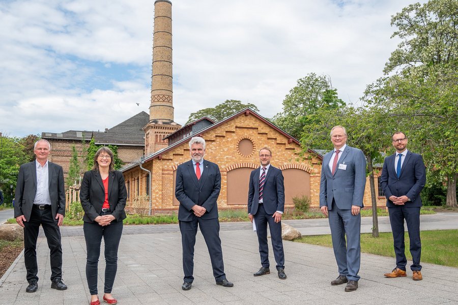 Fünf Männer und eine Frau stehen vor einem historischen Gebäude aus Backstein. Im Hintergrund ist ein Schornstein zu sehen.  Fünf Männer und eine Frau stehen vor einem historischen Gebäude aus Backstein. Im Hintergrund ist ein Schornstein zu sehen.