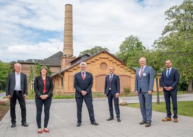 Fünf Männer und eine Frau stehen vor einem historischen Gebäude aus Backstein. Im Hintergrund ist ein Schornstein zu sehen.