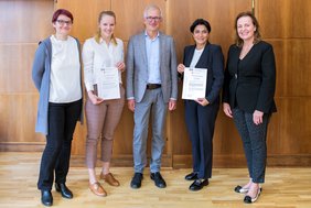 Fünf Personen stehen nebeneinander in einem holzvertäfelten Raum und lächeln in die Kamera. Zwei Frauen halten eine Urkunde in den Händen. 