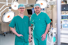 Zwei Personen in OP-Kleidung stehen in einem Operationssaal. Im Hintergrund und rechts im Bild befinden sich Lampen und andere OP-typische Geräte.