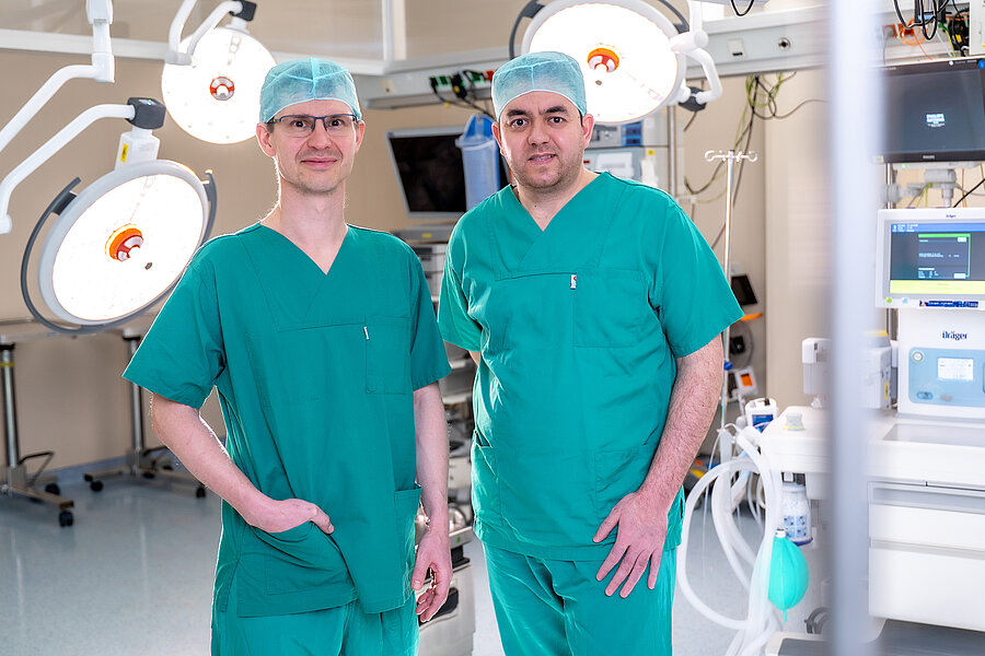 Zwei Personen in OP-Kleidung stehen in einem Operationssaal. Im Hintergrund und rechts im Bild befinden sich Lampen und andere OP-typische Geräte.  Zwei Personen in OP-Kleidung stehen in einem Operationssaal. Im Hintergrund und rechts im Bild befinden sich Lampen und andere OP-typische Geräte.