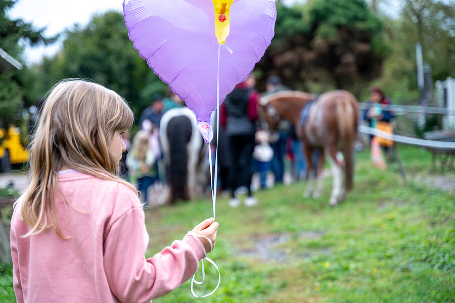 Ein Kind mit blonden Haaren und rosa Pullover steht im Vordergrund und hält einen lilafarbenen Luftballon in Herzform. Im Hintergrund sind mehrere Menschen und zwei Ponys zu sehen, die auf einer Wiese stehen.  Ein Kind mit blonden Haaren und rosa Pullover steht im Vordergrund und hält einen lilafarbenen Luftballon in Herzform. Im Hintergrund sind mehrere Menschen und zwei Ponys zu sehen, die auf einer Wiese stehen.