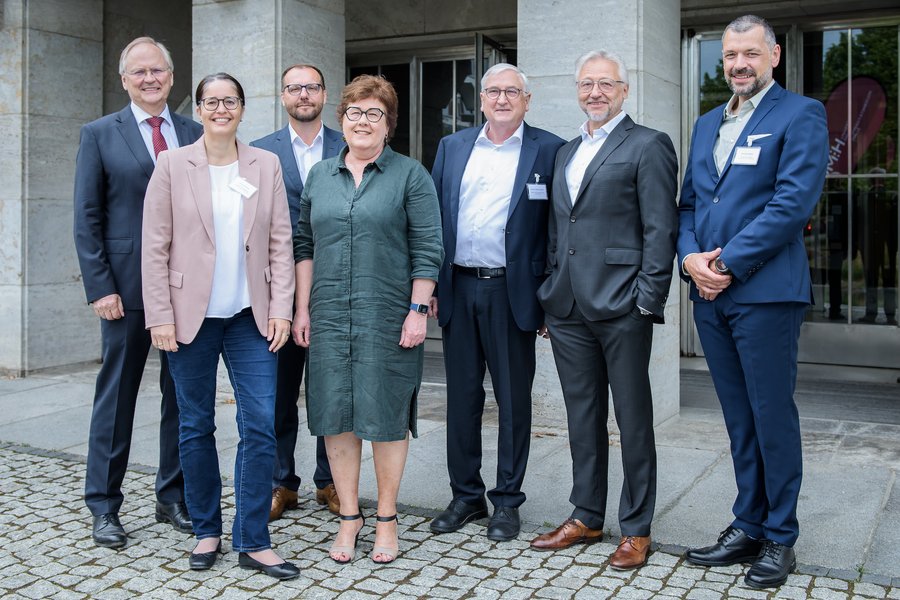 Zwei Frauen und fünf Männer stehen in Businesskleidung nebeneinander vor dem Eingang eines großen Gebäudes. Sie blicken lächelnd in die Kamera  Zwei Frauen und fünf Männer stehen in Businesskleidung nebeneinander vor dem Eingang eines großen Gebäudes. Sie blicken lächelnd in die Kamera