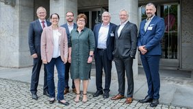 Zwei Frauen und fünf Männer stehen in Businesskleidung nebeneinander vor dem Eingang eines großen Gebäudes. Sie blicken lächelnd in die Kamera