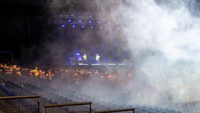  A large concert hall with dim light, a stage with people standing on it, white fog coming into the picture from the right.