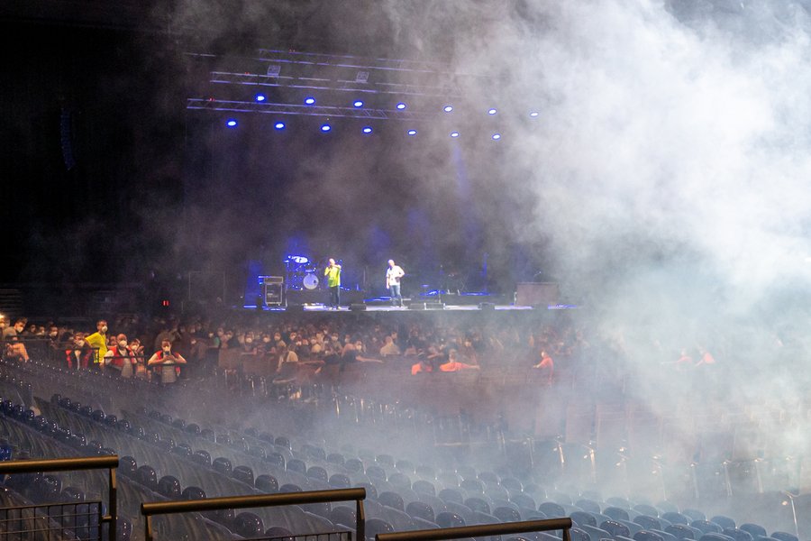 Ein großer Konzertsaal mit schummrigem Licht, einer Bühne, auf der Menschen stehen, von rechts kommt weißer Nebel ins Bild.  Ein großer Konzertsaal mit schummrigem Licht, einer Bühne, auf der Menschen stehen, von rechts kommt weißer Nebel ins Bild.
