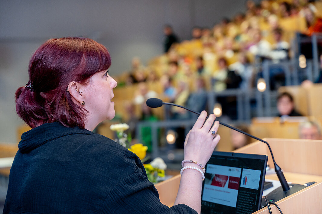 Eine Person steht in einem Hörsaal der Universitätsmedizin Halle und spricht in ein Mikrofon. In der Unschärfe befinden sich die Teilnehmenden des Geriatrietags 2026.