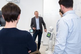 Menschen stehen mit dem Rücken zur Kamera. Durch eine Lücke sieht man in der Mitte einen Mann stehen, der gerade an einem Pepper-Roboter etwas erklärt.