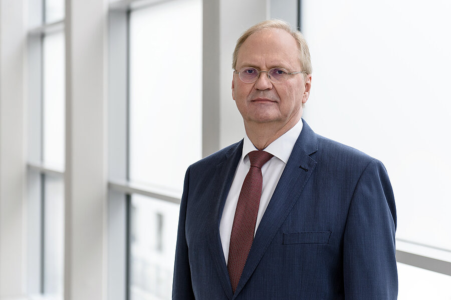 Älterer Mann mit grauem Haar, Brille und dunkelblauem Anzug im Portrait vor einer Fensterfront  Älterer Mann mit grauem Haar, Brille und dunkelblauem Anzug im Portrait vor einer Fensterfront