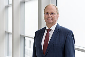 Älterer Mann mit grauem Haar, Brille und dunkelblauem Anzug im Portrait vor einer Fensterfront