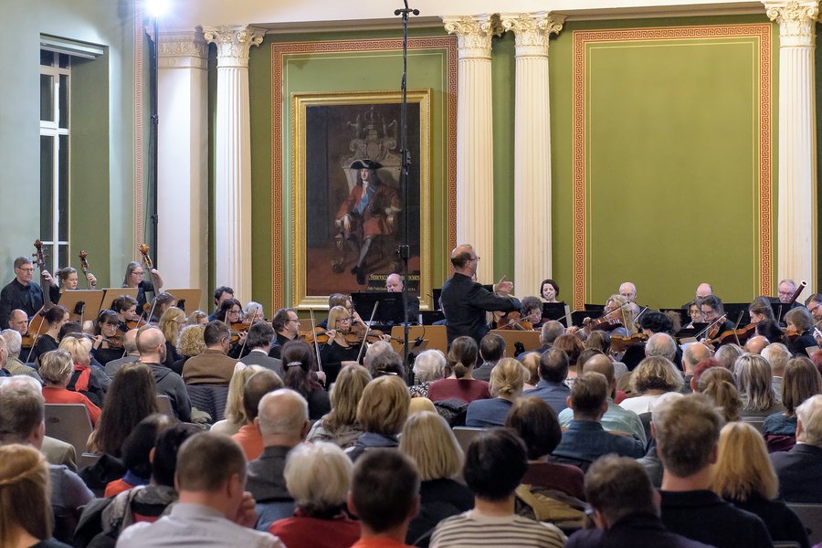 Orchester bei einem Konzert aus Zuschauerperspektive, in der Mitte stehend der Dirigent mit dem Rücken zum Publikum  Orchester bei einem Konzert aus Zuschauerperspektive, in der Mitte stehend der Dirigent mit dem Rücken zum Publikum