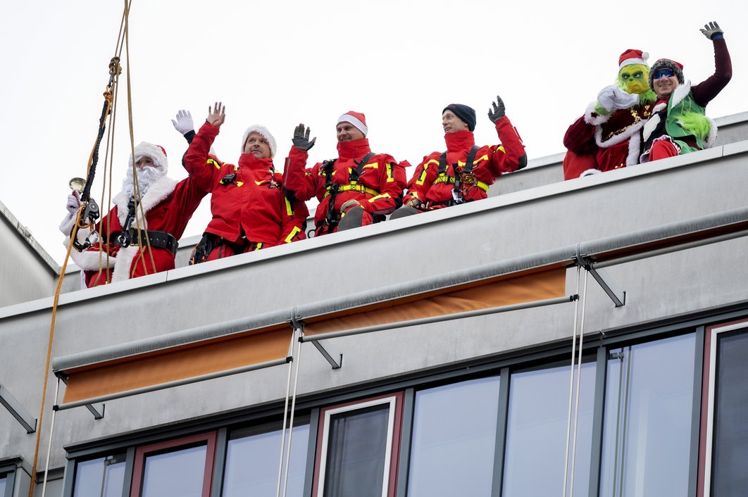Die Mitarbeiter der Feuerwehr Halle stehen auf dem Dach des Universitätsklinikums Halle (Saale) und winken. Einige Personen sind als Weihnachtsmann, Grinch und Wichtel verkleidet.