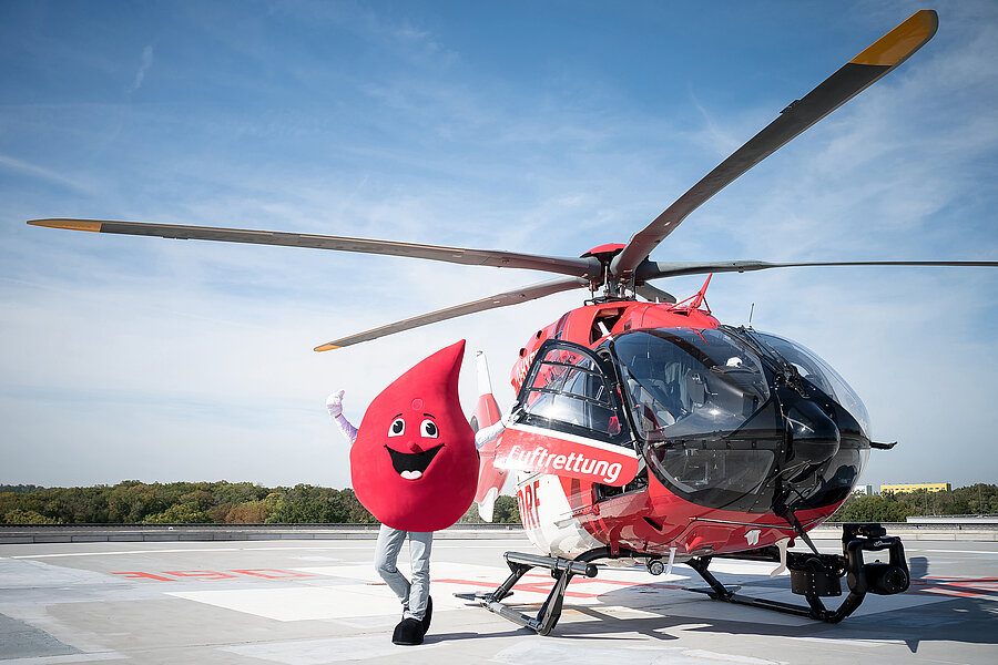 Ein Rettungshubschrauber steht auf dem Hubschrauberlandeplatz des Universitätsklinikums Halle (Saale). Daneben steht das Maskottchen der Blutspende und streckt die Arme in die Höhe.  Ein Rettungshubschrauber steht auf dem Hubschrauberlandeplatz des Universitätsklinikums Halle (Saale). Daneben steht das Maskottchen der Blutspende und streckt die Arme in die Höhe.