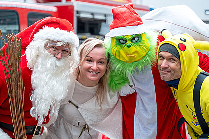 Eine Frau steht lächelnd zwischen drei verkleideten Personen: einem Weihnachtsmann, einem Grinch und jemandem im Pikachu-Kostüm.