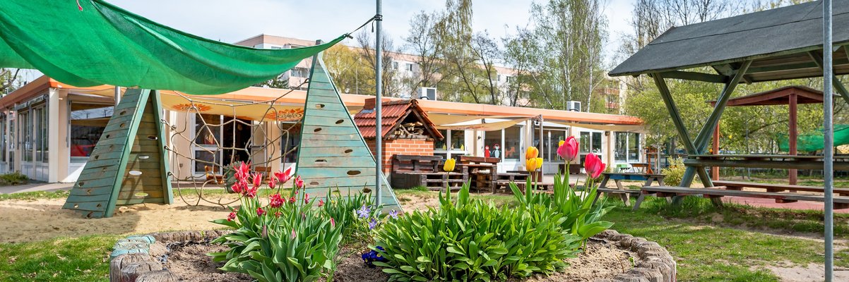 Blick auf den Spielplatz der Kita. Im Hintergrund ist das Gebäude zu sehen. Davor sind Blumen und ein Kletternetz für die Kinder.