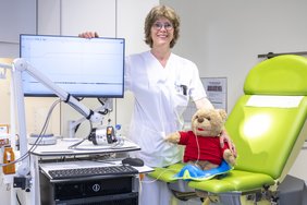 Eine Frau in medizinischer Arbeitskleidung steht zwischen einem medizinischen Messgerät mit Monitor und einem Behandlungsstuhl. Auf dem Stuhl sitzt ein Stofftier.