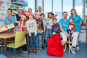 Gruppenbild von einer Gruppe Kinder und sechs Erwachsenen. Die Gruppe befindet sich im Spielzimmer einer Kinderstation eines Krankenhauses. Einige Personen tragen Weihnachtsmützen und halten Geschenke in den Händen. Ein Kind hält einen großen roten Sack mit Geschenken fest. 
