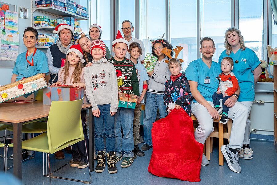 Gruppenbild von einer Gruppe Kinder und sechs Erwachsenen. Die Gruppe befindet sich im Spielzimmer einer Kinderstation eines Krankenhauses. Einige Personen tragen Weihnachtsmützen und halten Geschenke in den Händen. Ein Kind hält einen großen roten Sack mit Geschenken fest.   Gruppenbild von einer Gruppe Kinder und sechs Erwachsenen. Die Gruppe befindet sich im Spielzimmer einer Kinderstation eines Krankenhauses. Einige Personen tragen Weihnachtsmützen und halten Geschenke in den Händen. Ein Kind hält einen großen roten Sack mit Geschenken fest. 