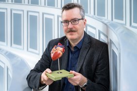 Ein Mann mit Brille, Schnauzbart und dunklem Anzug sitzt in historischen Hörsaal der halleschen Anatomie. Er hält mit beiden Händen ein Modell eines menschlichen Herzens in die Kamera.